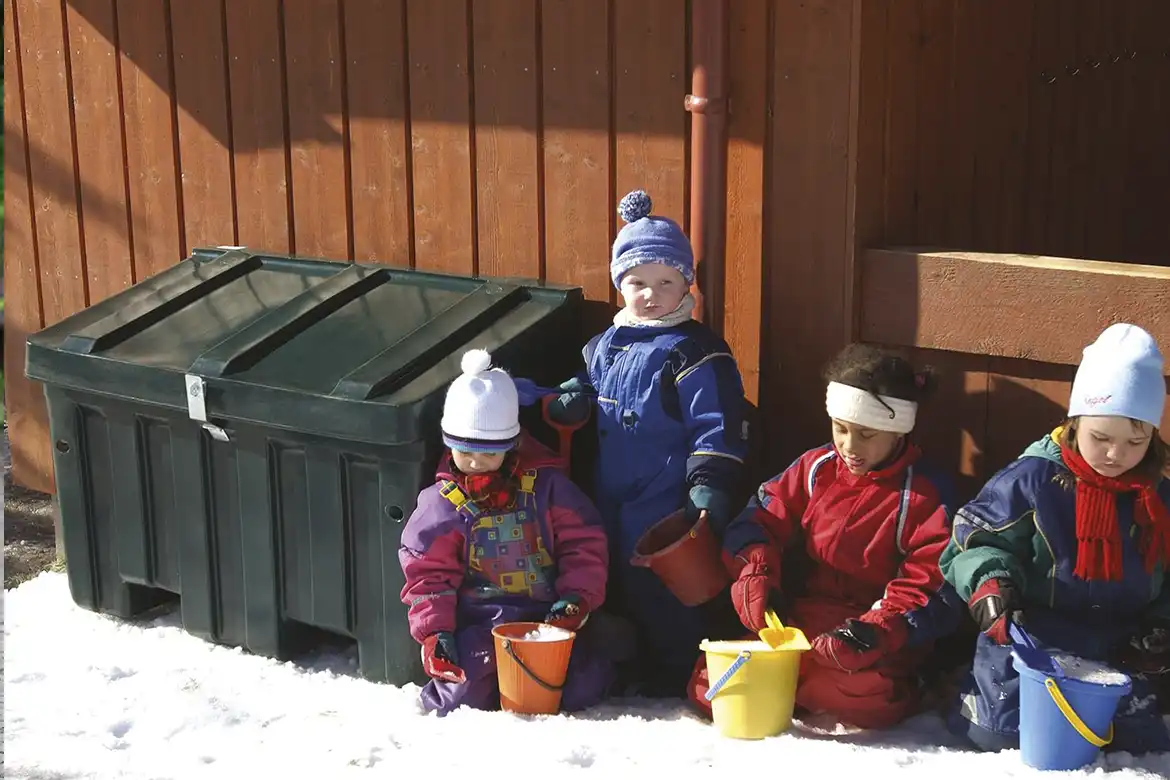 Fire små barn leker i snøen med spann og spade, de sitter foran en grønn oppbevaringskasse