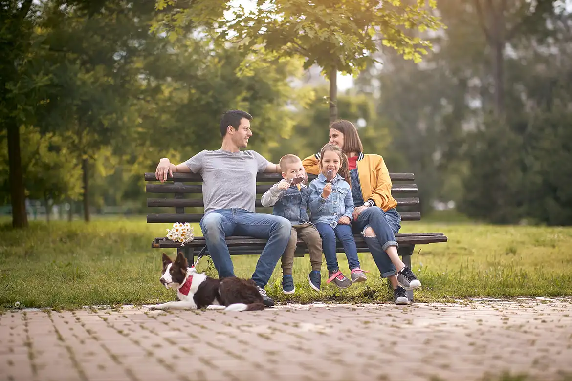 En familie på fire sitter på en benk i parken, de har med seg sin hund.